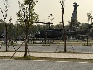 Một chiếc trực thăng quân sự màu xanh đậm được trưng bày ngoài trời trong một công viên hoặc bảo tàng. Ở phía sau, có xác của một chiếc máy bay khác được dựng đứng như một di tích. Xung quanh có nhiều cây cối và một vài người đang đi tham quan. Con đường ở phía trước được lát gạch và trải nhựa.。（日本語訳）。濃い緑色の軍用ヘリコプターが、公園か博物館の屋外に展示されています。後方には、別の飛行機の残骸が記念碑のように立てられています。周りにはたくさんの木があり、数人が見学しています。手前の道はレンガとアスファルトで舗装されています。