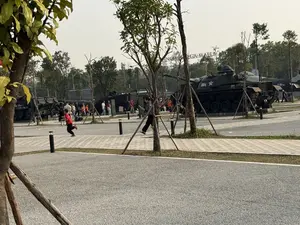 Bức ảnh chụp một không gian trưng bày ngoài trời, có nhiều người đang tham quan. Ở đây trưng bày các phương tiện quân sự cũ, bao gồm nhiều xe tăng và máy bay. Một chiếc xe tăng màu xanh đậm có ghi số "004" và một biểu tượng ngôi sao. Phía sau, một chiếc máy bay có chữ "FORCE" trên thân. Nhiều người lớn và trẻ em đang đi dạo xung quanh. Nổi bật có một cậu bé mặc áo phông đỏ đang chạy và một người phụ nữ đang đi bộ trong khi nhìn vào điện thoại. Khung cảnh có nhiều cây xanh, lối đi bộ lát gạch. Ở phía xa phía sau hàng cây có thể thấy một tấm biển lớn với dòng chữ "MEGA MALL".。（日本語訳）。写真は屋外の展示スペースを写しており、多くの人々が見学しています。ここでは、多くの戦車や航空機を含む古い軍用車両が展示されています。濃い緑色の戦車には「004」という数字と星のシンボルが書かれています。後方には、胴体に「FORCE」と書かれた航空機があります。多くの大人や子供が周りを散策しています。目立つのは、走っている赤いTシャツの少年と、携帯電話を見ながら歩いている女性です。風景には多くの緑の木々、レンガ敷きの歩道があります。木々の後ろの遠くに、「MEGA MALL」という文字の入った大きな看板が見えます。