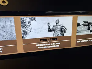 Màn hình trưng bày các giai đoạn lịch sử. Ở giữa là mốc thời gian 1788 - 1789 với chú thích bằng tiếng Anh 'RESISTANCE AGAINST THE QING DYNASTY' (Cuộc kháng chiến chống nhà Thanh) và hình ảnh đen trắng một bức tượng võ tướng. Bên trái là một phần hình ảnh về một trận thủy chiến. Bên phải là hình ảnh một cổng thành với chú thích 'THE NGUYEN DYNASTY' (Nhà Nguyễn).。（日本語訳）。歴史の各時代を展示するスクリーン。中央は、英語で「RESISTANCE AGAINST THE QING DYNASTY」（対清抗戦）と注釈された1788年～1789年の年表と、ある武将の像の白黒写真です。左側は、ある水戦の画像の一部です。右側は、「THE NGUYEN DYNASTY」（阮朝）と注釈された城門の画像です。