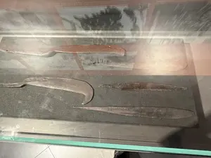 Bên trong một tủ kính trưng bày, có bốn công cụ bằng kim loại cổ, đã bị gỉ sét, được đặt trên một bề mặt tối màu. Ở phía trên cùng là một con dao lớn có hình dạng giống dao phay. Ở giữa là một công cụ hình lưỡi liềm và một vật nhọn giống mũi giáo. Ở dưới cùng là một lưỡi dao dài và nhọn. Tất cả các hiện vật đều có vẻ là đồ cổ, có thể là vũ khí hoặc nông cụ từ thời xưa.。（日本語訳）。展示用のガラスケースの中に、錆びついた4つの古い金属製の道具が、暗い色の表面に置かれています。一番上には、肉切り包丁のような形をした大きなナイフがあります。真ん中には、鎌の形をした道具と槍の穂先のような尖った物があります。一番下には、長くて尖った刃があります。すべての遺物は骨董品のようで、昔の武器か農具かもしれません。