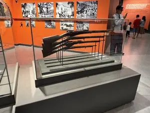 Bên trong một không gian bảo tàng, có một tủ kính trưng bày bốn khẩu súng trường cũ. Các khẩu súng được đặt trên giá đỡ như những hiện vật lịch sử. Phông nền là một bức tường màu cam sáng, trên đó có treo nhiều bức ảnh tư liệu đen trắng về các sự kiện lịch sử. Một số khách tham quan đang có mặt trong phòng. Một người đàn ông đội mũ, đeo kính đang đứng gần tủ kính và nhìn vào điện thoại của mình. Ở phía sau, một nhóm người khác bao gồm một người đàn ông mặc áo đỏ, một phụ nữ và một đứa trẻ đang xem các khu trưng bày khác.。（日本語訳）。博物館の空間内には、古いライフル銃4丁を展示するガラスケースがあります。銃は歴史的展示物として台座の上に置かれています。背景は明るいオレンジ色の壁で、そこには歴史的事件に関する多くの白黒の資料写真が掛けられています。何人かの来館者が室内にいます。帽子をかぶり眼鏡をかけた男性がガラスケースの近くに立ち、自分の携帯電話を見ています。後方では、赤いシャツを着た男性、女性、子供を含む別のグループが、他の展示エリアを見ています。