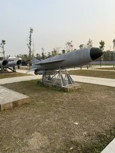 Một quả tên lửa lớn màu xám được trưng bày ngoài trời trên một bệ đỡ bằng kim loại. Tên lửa có phần mũi màu đen và các cánh ổn định ở phía đuôi. Bối cảnh là một khu vực triển lãm với các lối đi lát gạch và bãi cỏ. Ở phía sau, có thể nhìn thấy các khí tài quân sự khác, bao gồm một chiếc xe tăng mang số 250, một chiếc máy bay vận tải có số hiệu 282, và một phương tiện khác có số 555. Xa xa là cây cối và một tháp ăng-ten cao. Bầu trời u ám.。（日本語訳）。大きな灰色のミサイルが、金属製の台座の上に屋外展示されている。ミサイルは先端部分が黒く、尾部には安定翼がある。背景は、タイル敷きの通路と芝生のある展示エリアである。遠くには木々と高いアンテナ塔がある。空は曇っている。