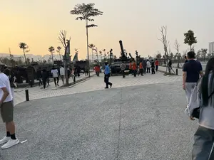 Bức ảnh chụp một khu trưng bày quân sự ngoài trời vào lúc hoàng hôn. Bầu trời có màu vàng cam, phía xa là các tòa nhà cao tầng. Nhiều người đang đi dạo và tham quan các hiện vật quân sự được trưng bày, bao gồm xe tăng, pháo tự hành và xác máy bay. Một nhân viên an ninh mặc đồng phục màu xanh đang đi tuần tra trong khu vực. Mọi người mặc trang phục thường ngày, đi lại trên con đường lát gạch và bãi đất rải sỏi.。（日本語訳）。写真は、夕暮れ時の屋外軍事展示エリアを写したものです。空はオレンジイエローで、遠くには高層ビルがあります。多くの人々が散歩し、戦車、自走砲、飛行機の残骸を含む、展示されている軍事展示物を見学しています。青い制服を着た一人の警備員がエリア内を巡回しています。人々は普段着で、レンガ敷きの道と砂利の地面の上を歩き回っています。