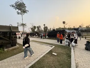 Một khung cảnh tại một khu trưng bày ngoài trời, có thể là bảo tàng quân sự, vào lúc hoàng hôn. Nhiều người, chủ yếu là giới trẻ, đang đi dạo và tham quan các hiện vật. Các hiện vật quân sự như xe tăng, xe bọc thép, pháo và máy bay được đặt trên bãi cỏ và dọc theo lối đi lát gạch. Ở phía trước, một cô gái mặc áo khoác đen và quần jean ống rộng đang đi bộ. Xa hơn, các nhóm người đang tụ tập, một số mặc áo màu cam, đang xem một chiếc xe bọc thép. Một vài người khác đang ngồi xổm trên lối đi để chụp ảnh. Bầu trời có màu vàng cam của buổi chiều tà, và có cây cối được trồng xen kẽ trong khu vực.。（日本語訳）。夕暮れ時の、屋外展示エリア、おそらく軍事博物館での光景。主に若者を中心とした多くの人々が、散策しながら展示物を見学している。手前では、黒いジャケットとワイドジーンズを履いた一人の少女が歩いている。さらに奥では、オレンジ色のシャツを着た人々を含むグループが集まり、一台の装甲車を見ている。他の数人は、写真を撮るために通路にしゃがみ込んでいる。空は夕暮れのオレンジがかった黄色で、エリア内には木々が点在して植えられている。