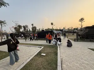 Khung cảnh một khu trưng bày ngoài trời, có thể là bảo tàng quân sự, vào buổi chiều tà. Nhiều loại khí tài quân sự như xe tăng, xe bọc thép, pháo và máy bay được đặt trên bãi cỏ. Rất đông người, chủ yếu là các bạn trẻ, đang đi dạo, tham quan và chụp ảnh xung quanh các hiện vật. Một số người mặc đồng phục áo màu cam. Lối đi được lát gạch xen kẽ với các khu vực trồng cỏ. Bầu trời trong xanh và có ánh vàng của hoàng hôn.。（日本語訳）。夕暮れ時の、おそらく軍事博物館であろう屋外展示エリアの光景。主に若者を中心とした多くの人々が、展示物の周りを散策したり、見学したり、写真を撮ったりしている。オレンジ色のシャツの制服を着ている人もいる。通路は、芝生が植えられたエリアと交互にタイルで舗装されている。空は澄み切った青色で、夕焼けの黄金色の光が差している。