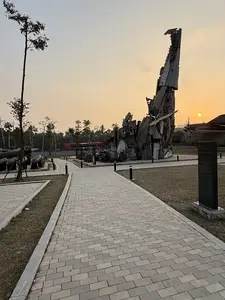 Khung cảnh một khu trưng bày ngoài trời vào lúc hoàng hôn. Ở trung tâm là một tác phẩm điêu khắc lớn làm từ xác máy bay bị phá hủy, dựng đứng như một đài tưởng niệm. Một lối đi bộ lát gạch xám chạy xuyên qua khu vực. Xung quanh có các hiện vật chiến tranh khác như bom, động cơ và một phần của một chiếc máy bay vận tải lớn ở phía sau. Bầu trời có màu cam ấm áp của buổi chiều tà với mặt trời đang lặn ở phía xa.。（日本語訳）。夕暮れ時の屋外展示エリアの光景。中央には、破壊された航空機の残骸から作られた大きな彫刻が、記念碑のように直立している。灰色のタイルが敷かれた歩道がそのエリアを貫いている。周囲には、爆弾、エンジン、そして後方には大型輸送機の一部など、他の戦争遺物がある。空は夕暮れの暖かいオレンジ色で、遠くには太陽が沈んでいる。