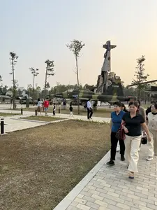 Bức ảnh ghi lại cảnh các du khách đang tham quan một bảo tàng ngoài trời, có khả năng là bảo tàng chứng tích chiến tranh. Nhiều máy bay quân sự, bao gồm máy bay chiến đấu và trực thăng được sơn rằn ri, đang được trưng bày trên bãi cỏ. Nổi bật ở phía sau là một công trình lớn được làm từ các mảnh vỡ của máy bay. Một số nhóm du khách, bao gồm cả người lớn, trẻ em và người cao tuổi, đang đi bộ trên lối đi lát gạch để quan sát các hiện vật. Khung cảnh diễn ra vào một buổi chiều trời quang, tạo nên một không gian tham quan yên bình.。（日本語訳）。写真は、観光客たちが屋外博物館を見学している光景を捉えている。おそらく戦争証跡博物館だろう。迷彩塗装された戦闘機やヘリコプターを含む多くの軍用機が、芝生の上に展示されている。後方には、飛行機の残骸から作られた大きな構造物が際立っている。大人、子供、高齢者を含むいくつかの観光客のグループが、展示物を観察するために舗装された通路を歩いている。光景は晴れた日の午後に繰り広げられており、平和な見学空間を作り出している。
