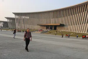 Một người đàn ông châu Á mặc áo sơ mi kẻ sọc, đeo ba lô đang đi bộ trên một quảng trường rộng lớn. Phía sau ông là một tòa nhà kiến trúc hiện đại, to lớn với mặt tiền được tạo thành từ nhiều cột dọc và có hình dạng cong. Có nhiều người khác đang đi lại và tụ tập ở phía xa, gần lối vào tòa nhà. Bầu trời có màu sáng, mờ ảo.。（日本語訳）。ストライプのシャツを着てバックパックを背負ったアジア人男性が、広大な広場を歩いている。彼の後ろには、多くの縦の柱で構成され、湾曲した形状のファサードを持つ、大きくて近代的な建物がある。遠くの建物の入り口付近では、他の多くの人々が行き来したり集まったりしている。空は明るく、霞んでいる。